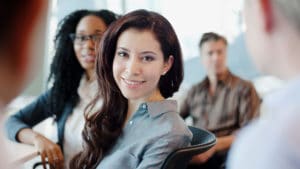 Woman labor relations specialist smiles at camera and sits around a table with a diverse group of employees in a work setting