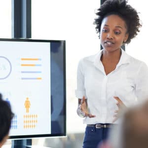 Woman management analyst in a white shirt stands in an office next to a computer monitor with graphs and charts and presents information to audience