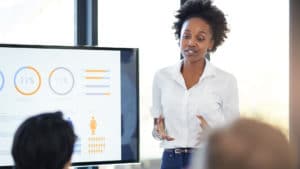 Woman management analyst in a white shirt stands in an office next to a computer monitor with graphs and charts and presents information to audience