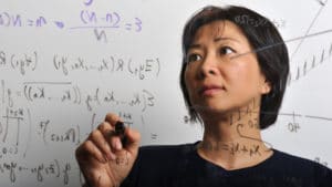 Female mathematician using a black marker writing formula on a clear piece of glass with another whiteboard behind her