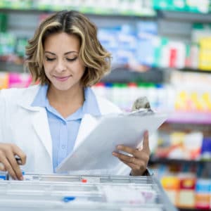 woman pharmacist in light blue shirt and white pharmacy coat stands in front of shelves with colorful medicines while she fills a prescription