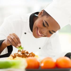 Female chef in a white coat and hat smiles while leaning over a special desert and adds a small piece of green fruit