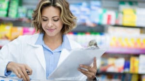 woman pharmacist in light blue shirt and white pharmacy coat stands in front of shelves with colorful medicines while she fills a prescription