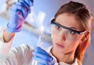 Chemistry College Major - Woman wearing lab coat, purple nitrile gloves and safety glasses uses a dropper to add a liquid into a test tube