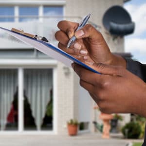 Woman real estate appraiser wearing a gray suit stands in front of a modern home with solar panel roof and holds a notebook