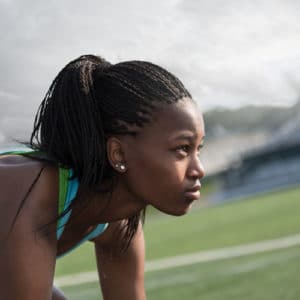Female track athlete runs in her lane on dark blue track in late afternoon next to a field with green grass