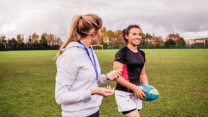 Female athletic trainer works with a woman athlete carrying a blue ball across a field with green grass and trees in background