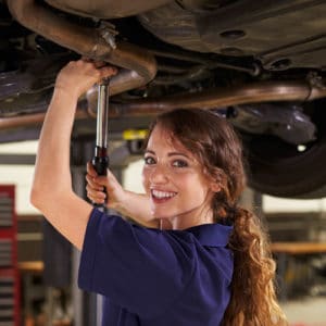 Female auto mechanic in dark blue work shirt and smiles while standing under a car and using a wrench to fix a car