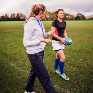 Female athletic trainer works with a woman athlete carrying a blue ball across a field with green grass and trees in background