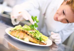 Culinary Arts College Major - Female chef in restaurant kitchen in white smock carefully places greens on a plate with salmon