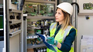 Female electrical engineer wearing a yellow safety vest and hard hat takes note while examining an electric panel