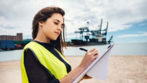 Female marine engineer in black shirt and yellow safety vest taking notes in front of ship while standing on a beach near an industrial harbor