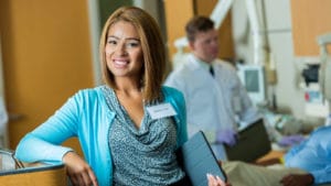 Woman medical manager at a physician's office wearing a bright blue sweater and holding a laptop and smiling towards the camera