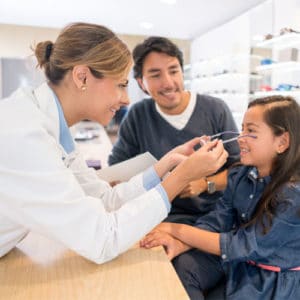 Woman optometrist in a light blue shirt and white doctor's coat places new glasses onto a young smiling girls face next to her father