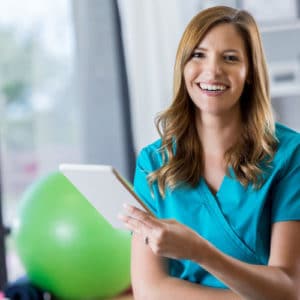 Female physical therapist in a blue uniform smiles at the camera and holds a green exercise ball in a treatment room with large window