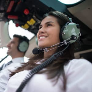 Female pilot in white uniform wearing light green headphones smiles as she sits with copilot in cockpit and flies plane
