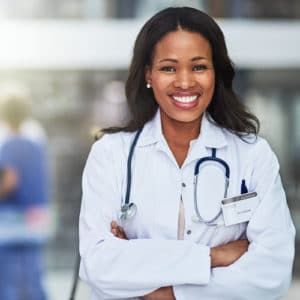 Woman doctor with a white coat and stethoscope around her collar stands smiling at the camera in a hospital with other physicians