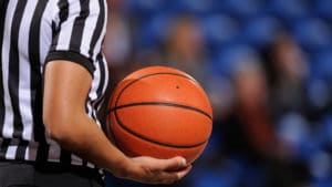Woman referee standing and wearing a black and white vertically striped ref shirt and holding a basketball in a sports arena