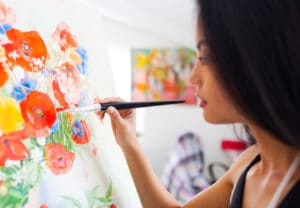 Woman paints brightly colored flowers on a white canvas placed on an artist easel