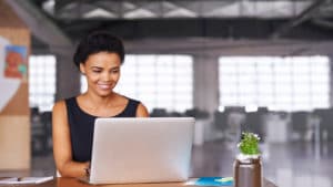 Woman web developer designs website on computer laptop sitting a table in a large loft office space with lots of windows