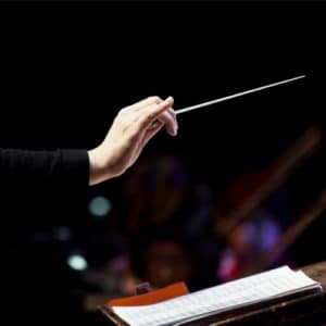 Woman conductor holds a baton above a podium to direct an orchestra playing from a composer's sheet music