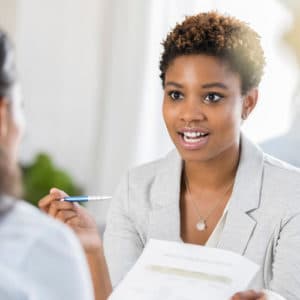 Woman insurance underwriter holds a notepad and pencil and speaks with a fellow worker to show her a question form.