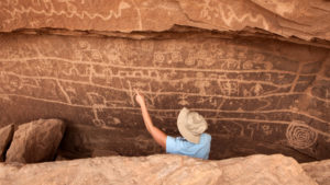 Anthropologist and archeologist in khaki hat studying ancient drawings on wall of a sandstone cave