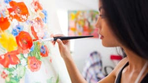 Female artist painting brightly colored red yellow and blue flowers and green grass on a canvas on easel