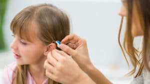 Female audiologist in white coat adjusts a hearing aid on a girl's ear while inside of a bright doctors office room