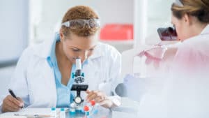 Woman microbiologist in white lab coat and safety glasses on her head examines molecules with a microscope in laboratory