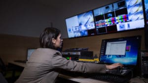 Female broadcast engineer working to adjust sound levels at audio mixer and computer with rows of monitors at live performance