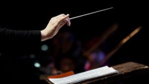 Woman conductor holds a baton above a podium to direct an orchestra playing from a composer's sheet music