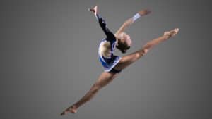 Female dancer in a white and blue outfit athletically leaps into the air on stage with toes pointed against a gray background