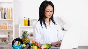 Female dietician in a white coat stands next to a table with a variety of fresh fruit and vegetables including a pineapple