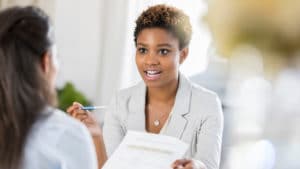 Woman insurance underwriter holds a notepad and pencil and speaks with a fellow worker to show her a question form.