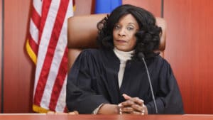 Woman judge sits at her desk wearing a black robe in a courtroom with a flag and wood paneled wall behind her brown chair