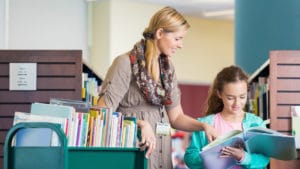 Woman librarian smiles and helps a girl find a book next to a green rolling cart in a colorful well lit library room