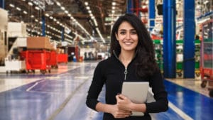 Woman logistician in a black shirt stands on a shiny gray concrete floor in a colorful manufacturing facility holding a notebook