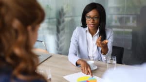 Female marketing manager in a light blue jacket sits at an office table and presents information from a colorful pie chart to fellow worker