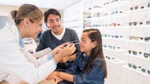 Woman optometrist in a light blue shirt and white doctor's coat places new glasses onto a young smiling girls face next to her father