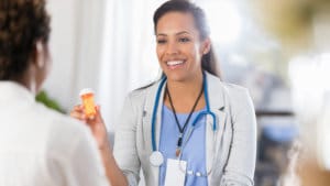 Woman psychiatrist in light blue medical scrubs and white coat smiles at a patient and shows a recommended prescription