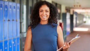 Woman school principal in a blue shirt stands in a school hallway next to shiny blue lockers and holding a clipboard