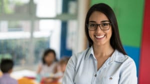 Woman teacher with glasses smiles and stands in front of a brightly painted green red and blue classroom of children
