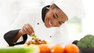 Female chef in a white coat and hat smiles while leaning over a special desert and adds a small piece of green fruit
