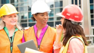 Women construction managers with orange safety vests and hard hats hold notebooks and converse at a work site