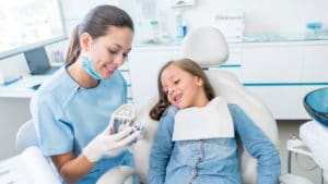 Female dentist in blue scrubs and white nitrile gloves shows a girl patient in dental chair a mold of her teeth