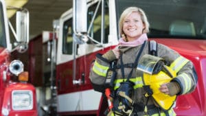 Female firefighter in protective safety coat and gloves stands in front of a red fire truck with white trim inside of a station