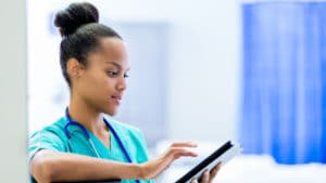 Woman nurse in bright green scrubs and stethoscope stands in a hospital room with blue curtains and takes notes on a tablet