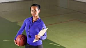 Female coach stands on an indoor multipurpose athletic court with a clipboard in one hand and a basketball in the other