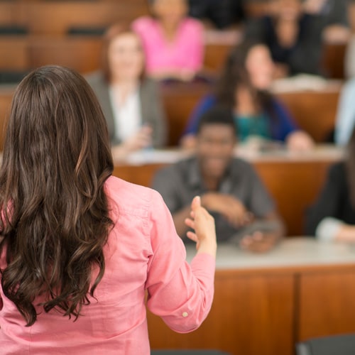 Female college professor stands in front of students in a university classroom with rows of wood grain front desks